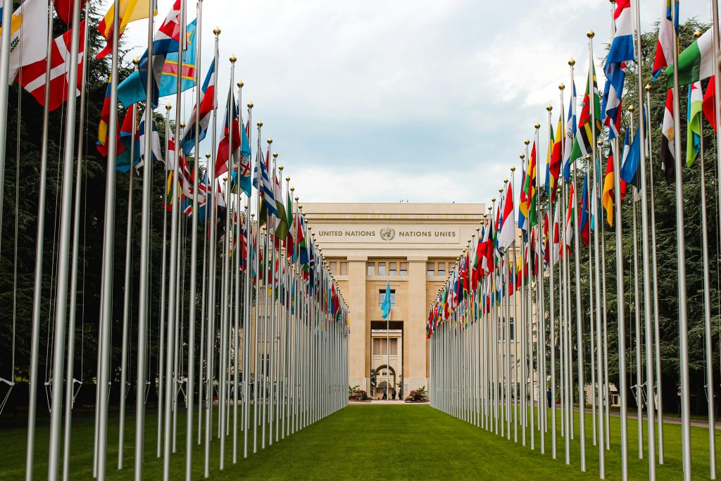 UN building in Geneva. An impressive, tan frontage bearing the UN logo is at the end of a double avenue of flagpoles, each bearing one of the national flags of the States Members of the UN