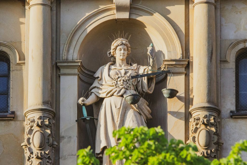 A statue of justice in Hamburg. A figure sits in an alcove, holding a set of discoloured copper and brass scales in her left hand, and her right hand resting on a sword. She looks towards the scales. She is flanked by two columns and the top branches of a green tree make up the foreground