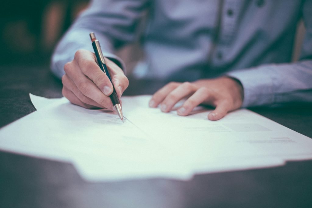 A man in a blue shirt sits at a dark wood desk. Only his waist to his chest and his hands are visible. He is holding a black ballpoint pen with gold details, and is writing on papers on the desk top.